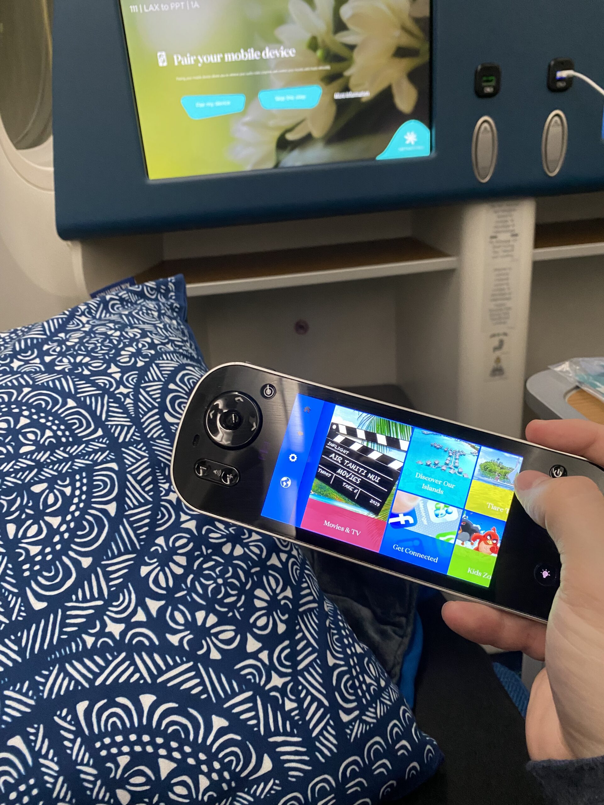 The Panasonic eX3 IFE controller in my hand — you can see the LAX to PPT flight info and the Air Tahiti Nui movie library on the handset screen